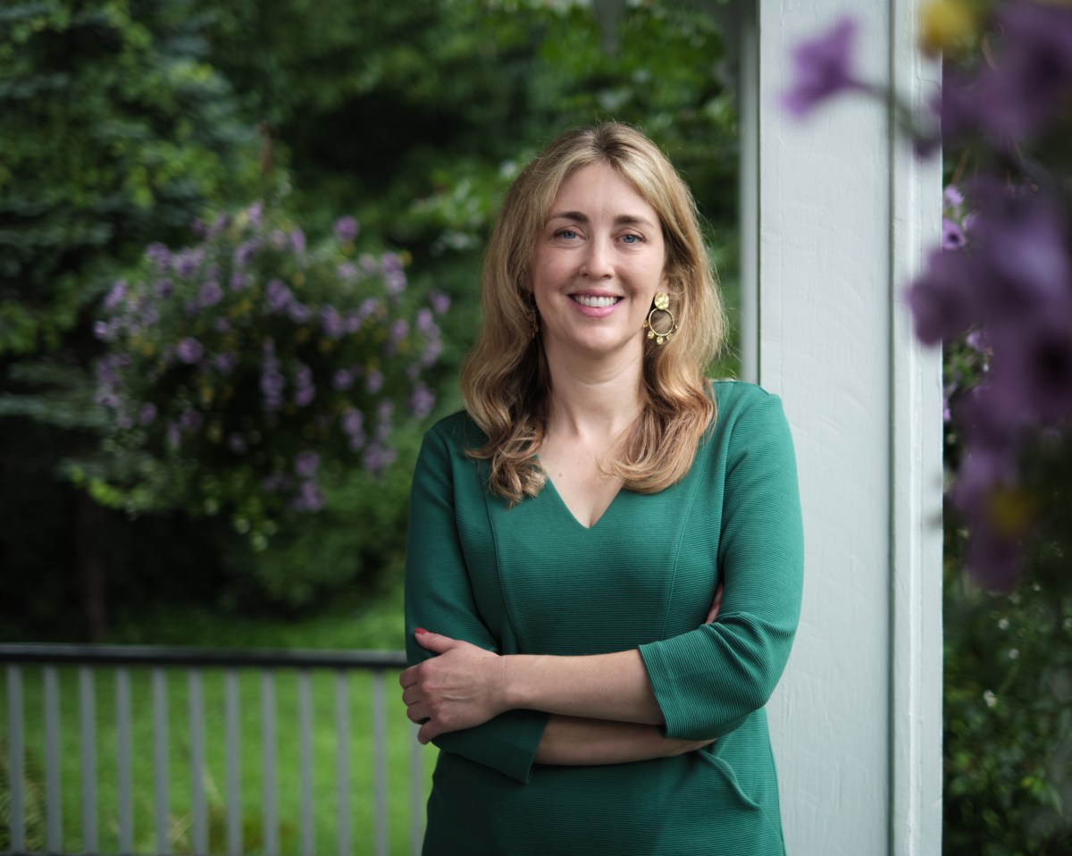 A woman with blonde hair in a green dress stands on a porch with her arms crossed, smiling, with greenery and purple flowers in the background.