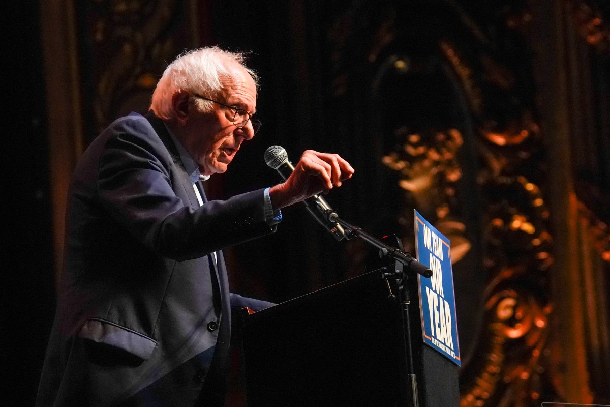 An older man in a suit speaks passionately at a podium with a microphone on a dimly lit stage; a partially visible sign reads "THE YEAR.