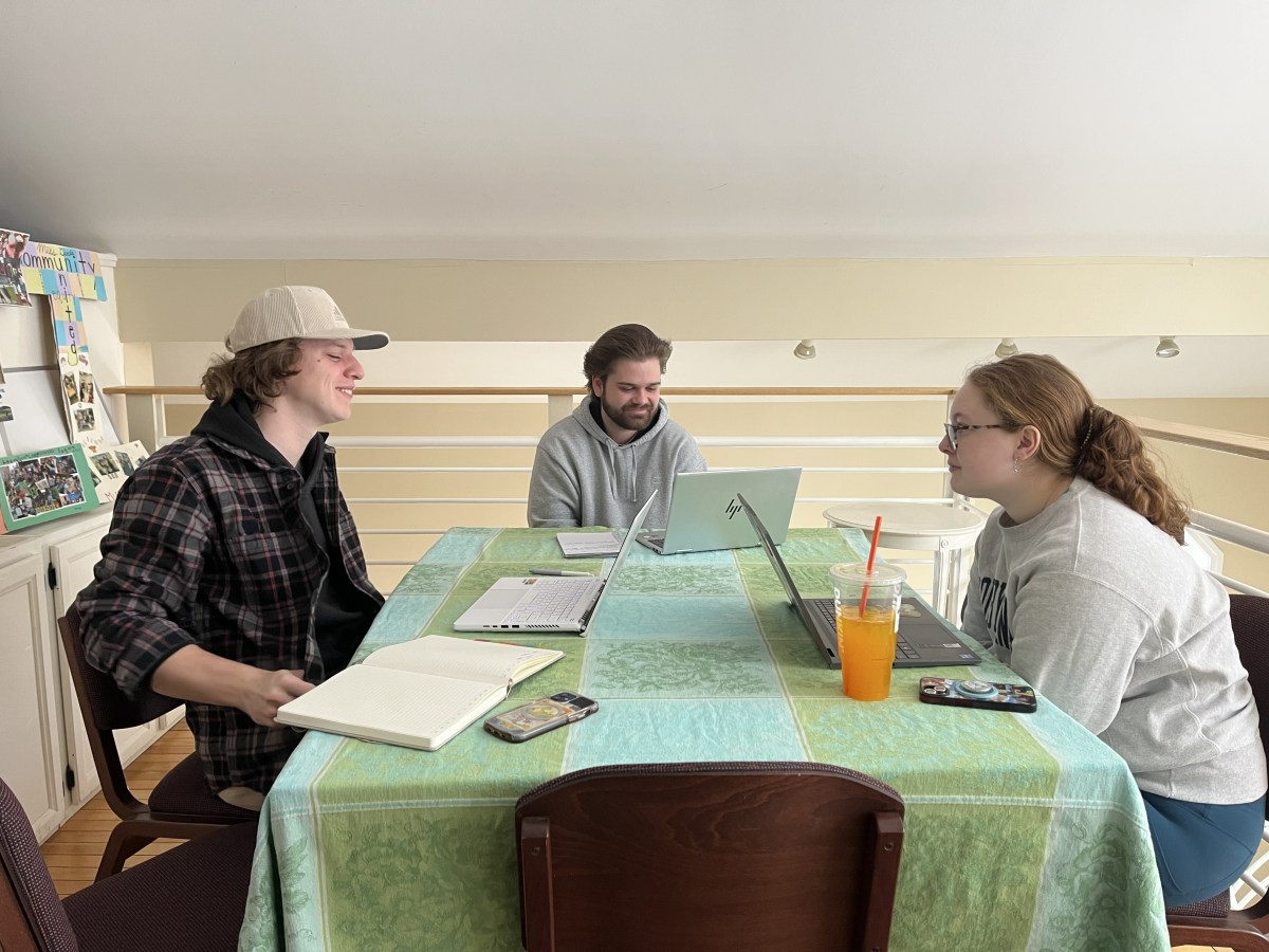 Three people sit around a table with laptops, notebooks, and a drink, appearing to work or study together in a well-lit indoor space.
