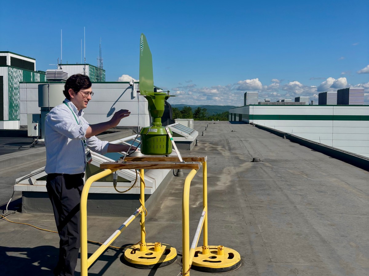 A person stands on a rooftop beside a small green wind turbine mounted on a yellow frame, with a laptop on the table and clear skies in the background.