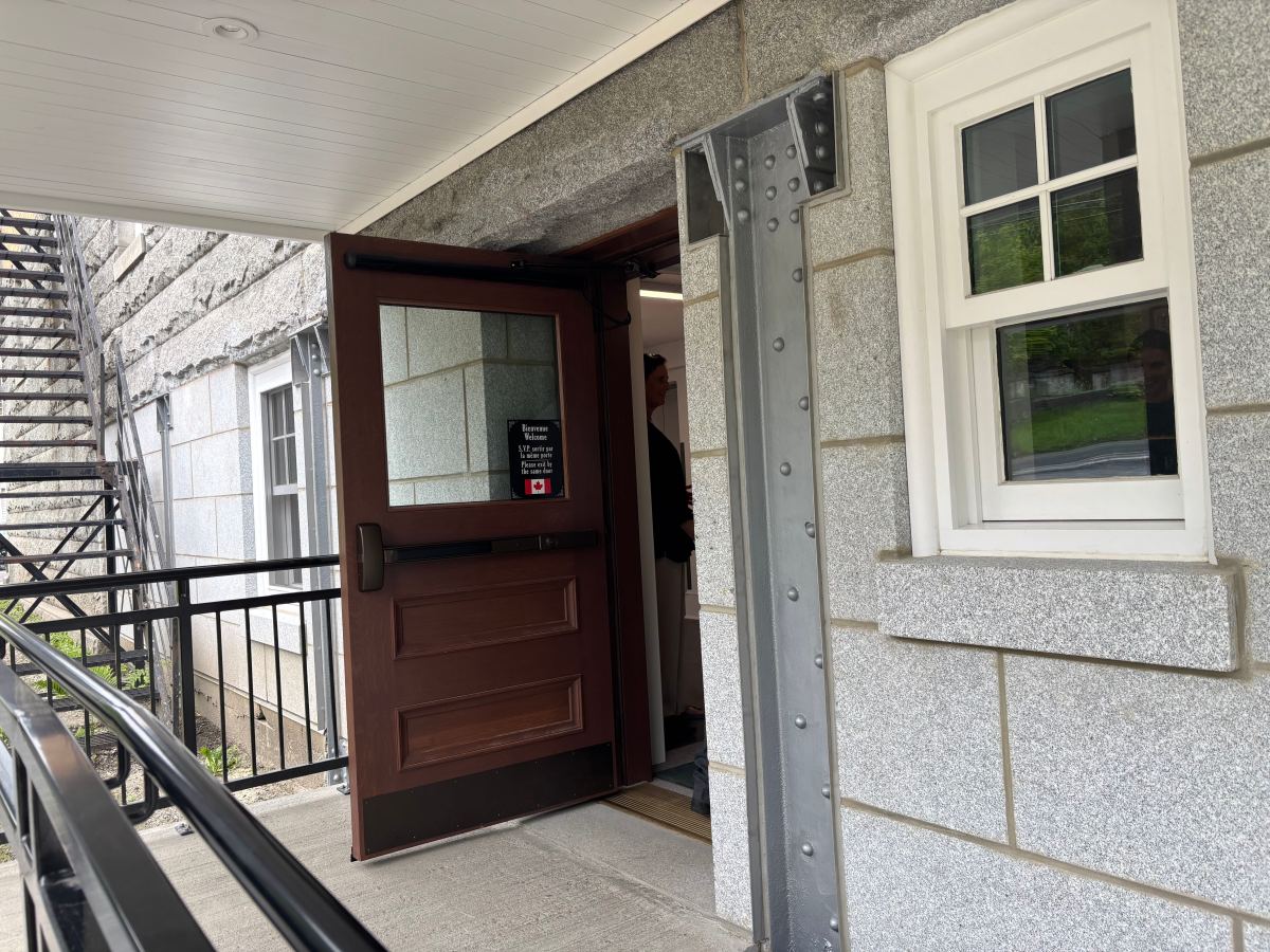 A brown door propped open at a stone building entrance with a metal ramp and railing; part of a staircase and window are visible.