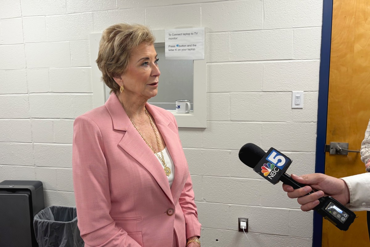 A woman in a pink blazer is being interviewed by someone holding an NBC 5 microphone in a room with white brick walls.