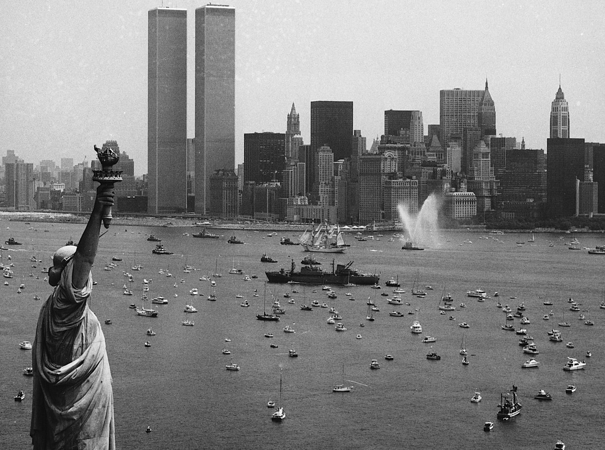 Black and white photo of the Statue of Liberty facing New York Harbor, with numerous boats and the World Trade Center towers visible in the Manhattan skyline.