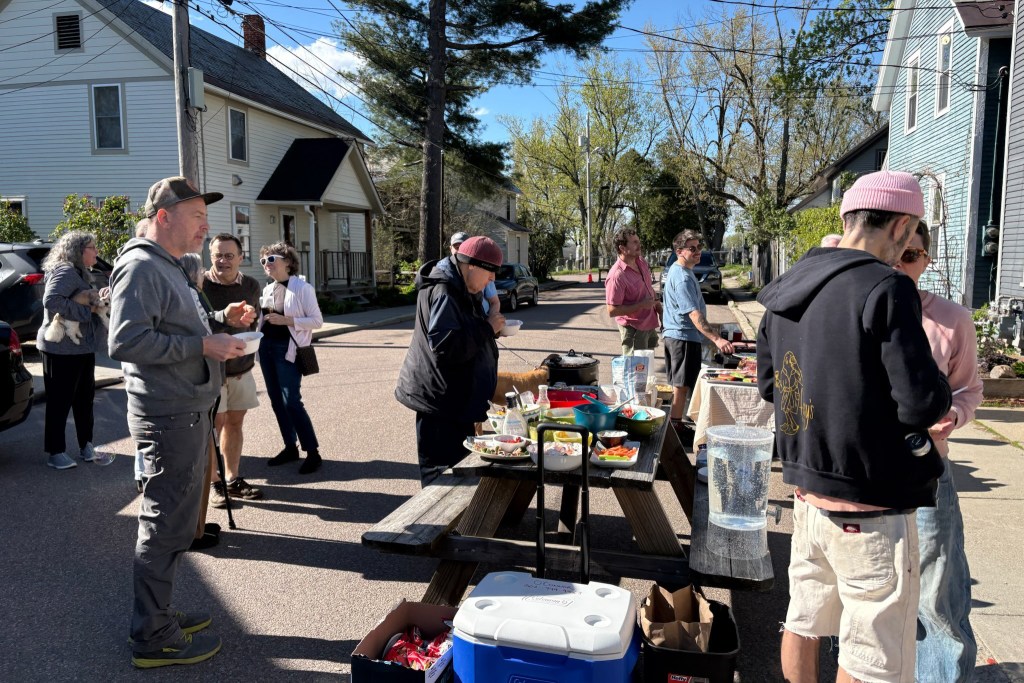 A group of people gather around picnic tables with food and drinks on a sunny residential street, with houses and trees in the background.