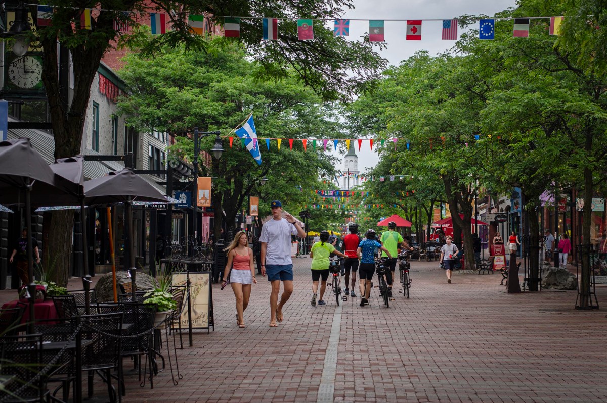 Pedestrian street lined with trees, outdoor seating, people walking and cycling, and strings of international flags overhead.