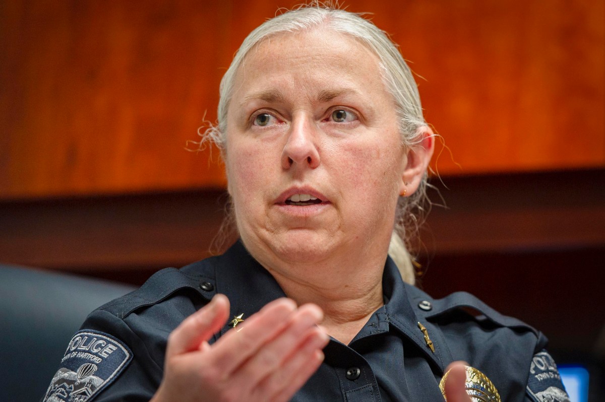 A police officer with light hair pulled back, wearing a uniform, sits indoors and gestures with one hand while speaking.