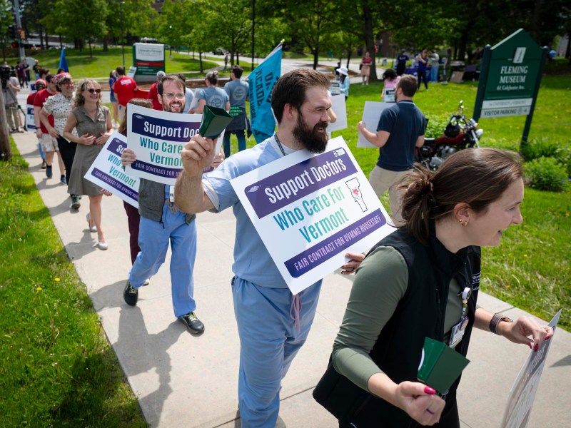 A group of people holding signs that read “Support Doctors Who Care for Vermont” walk in a peaceful protest along a sidewalk.