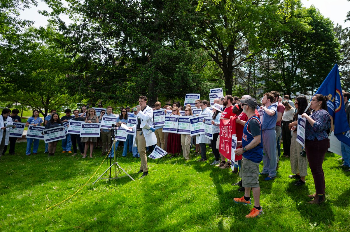 A group of people stand on grass holding signs while one person speaks into a megaphone at a public outdoor gathering.