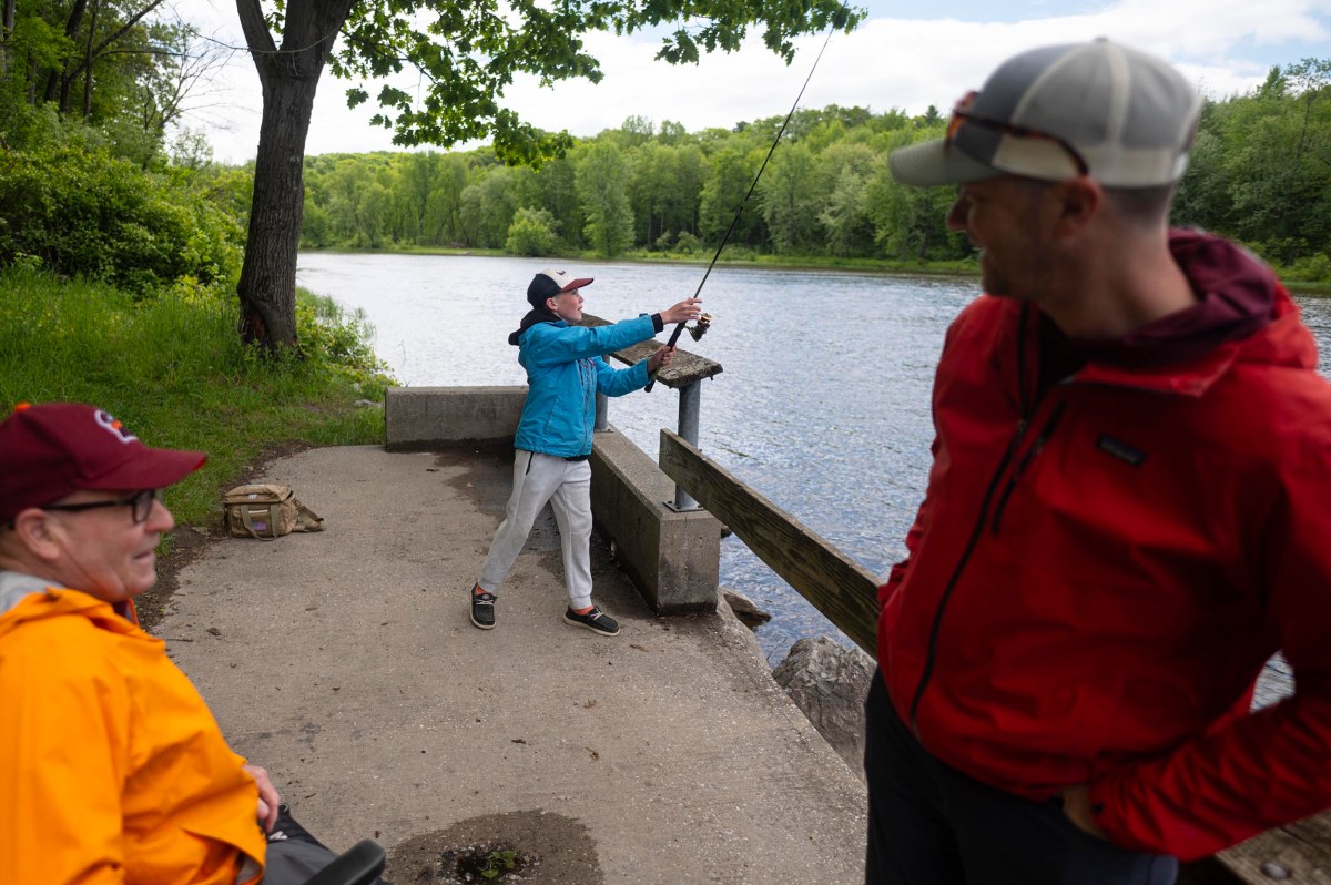 A child in a blue jacket casts a fishing line by a river while two adults watch nearby; the scene is outdoors with green trees in the background.