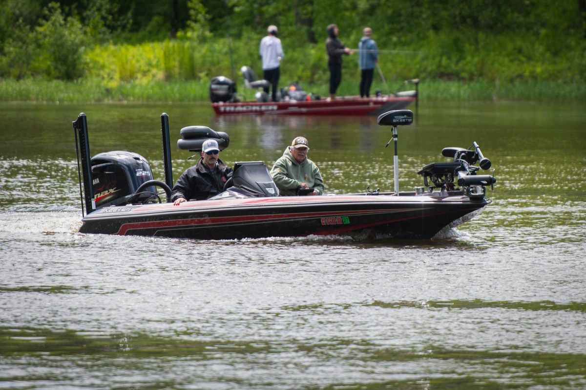 Two people ride in a black bass fishing boat on a river, with another red boat and three people visible in the background near the shore.