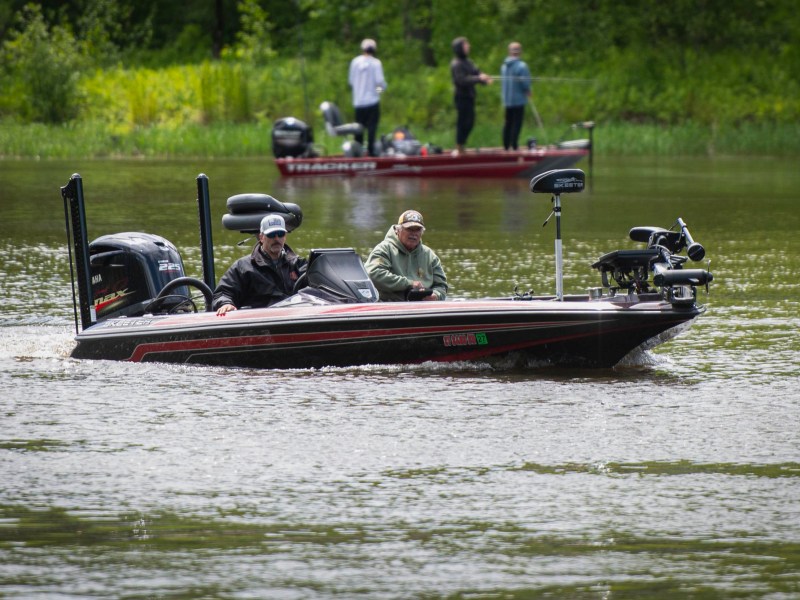 Two people ride in a black bass fishing boat on a river, with another red boat and three people visible in the background near the shore.
