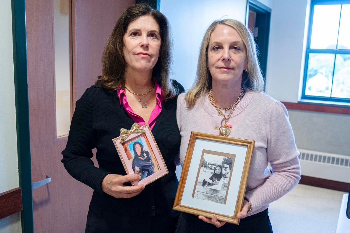 Two women stand indoors holding framed photographs of other women or girls. They face the camera with neutral expressions.