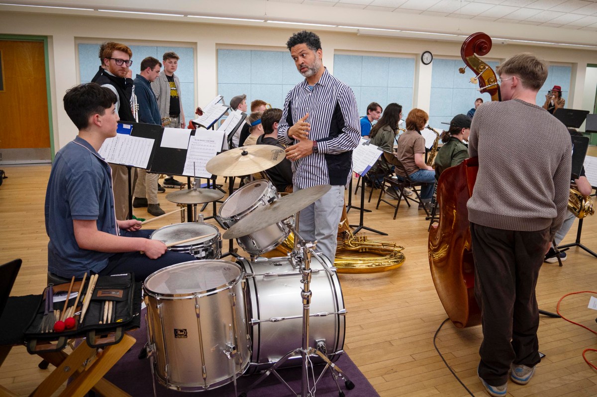 A music instructor speaks to a student at a drum set while other students rehearse with various instruments in a classroom.