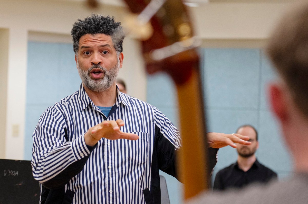 A man in a striped shirt gestures with his hands while speaking, with a blurred person and a musical instrument visible in the foreground.