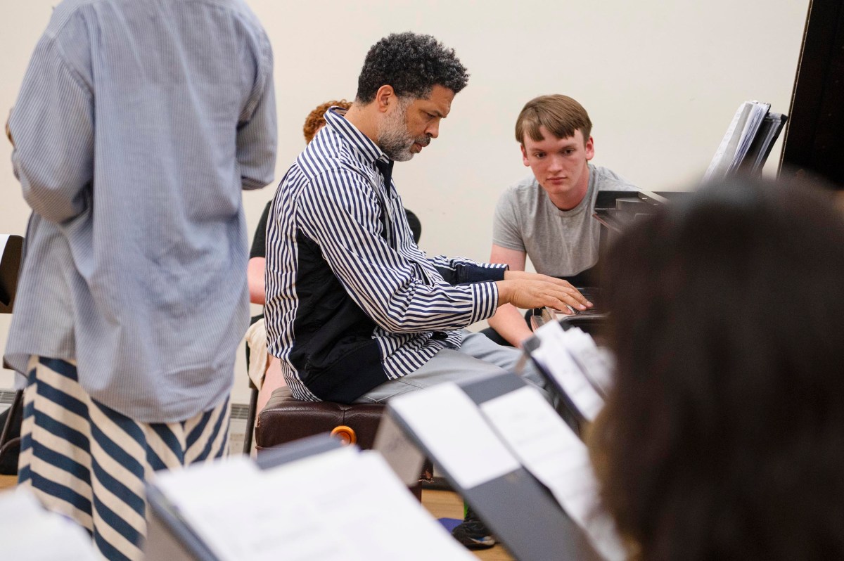 A man plays the piano while a young man watches closely; other people are present in the background of the room.