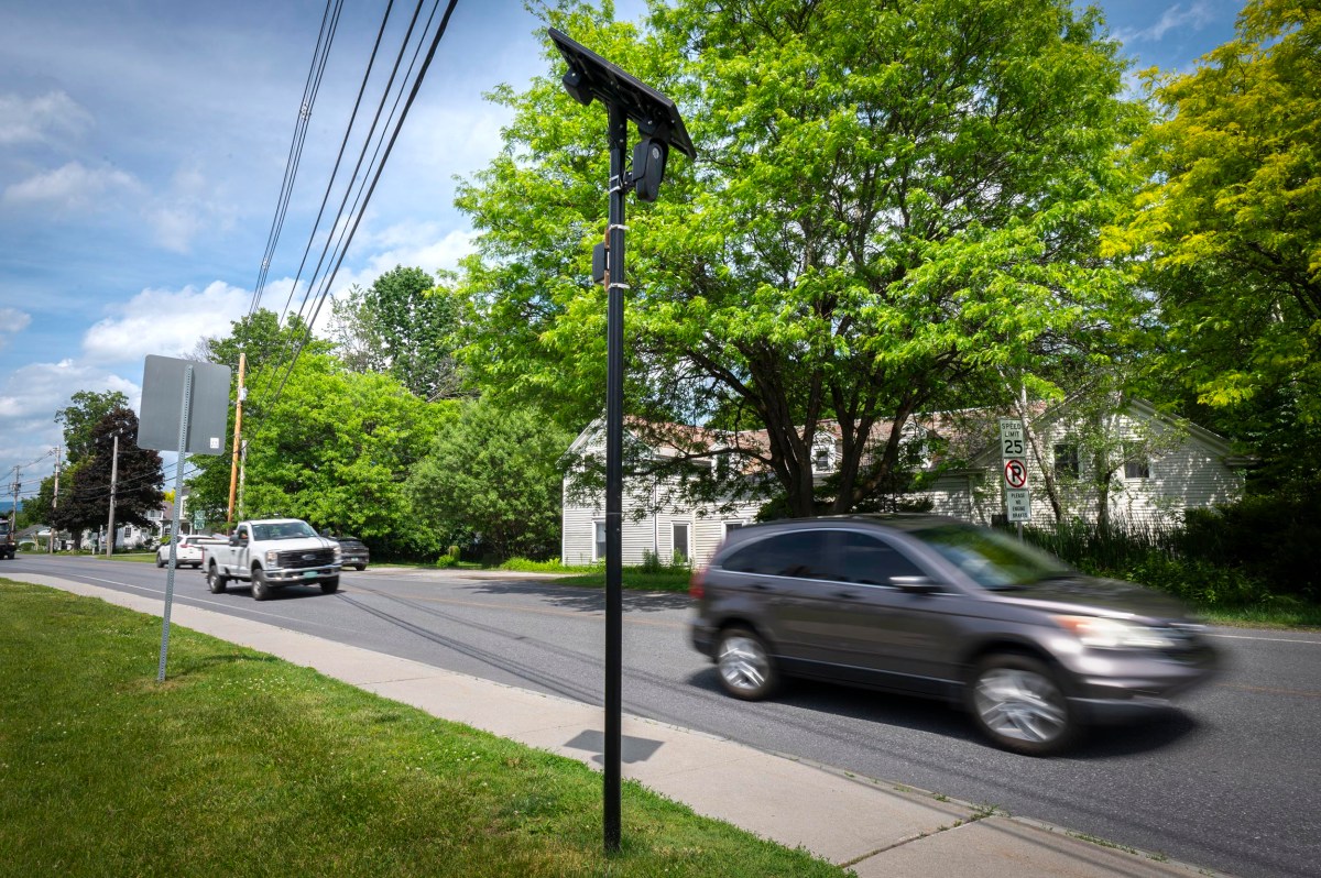 Cars drive past a solar-powered speed monitoring sign on a suburban street lined with trees and houses.