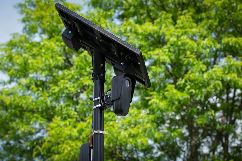 A black security camera and solar panel mounted on a pole, with green trees in the background.