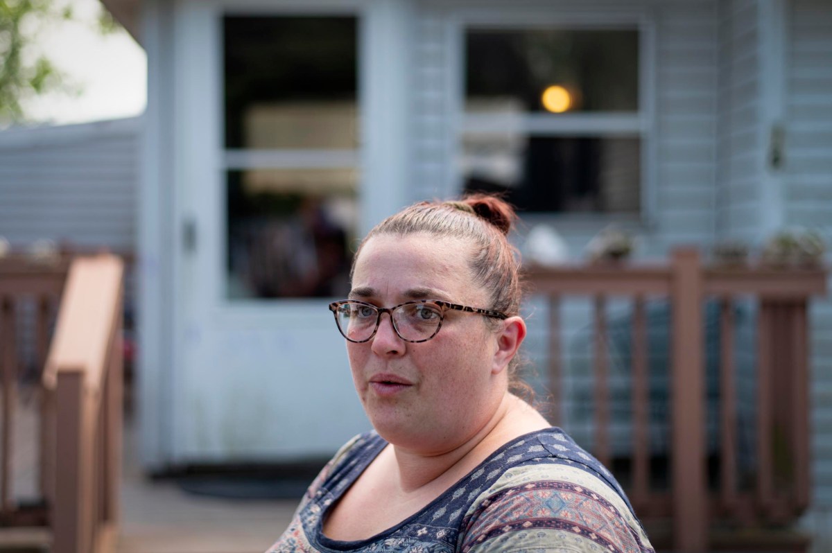 A woman with glasses and a patterned shirt sits outdoors in front of a house with a wooden porch and a glass door.