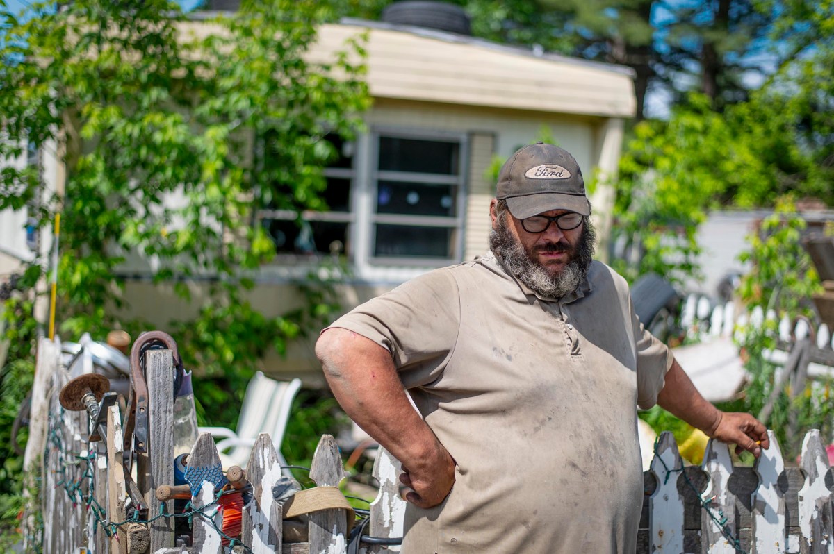 A man in a Ford cap stands behind a white picket fence with garden tools; a house with peeling paint and broken windows is in the background.