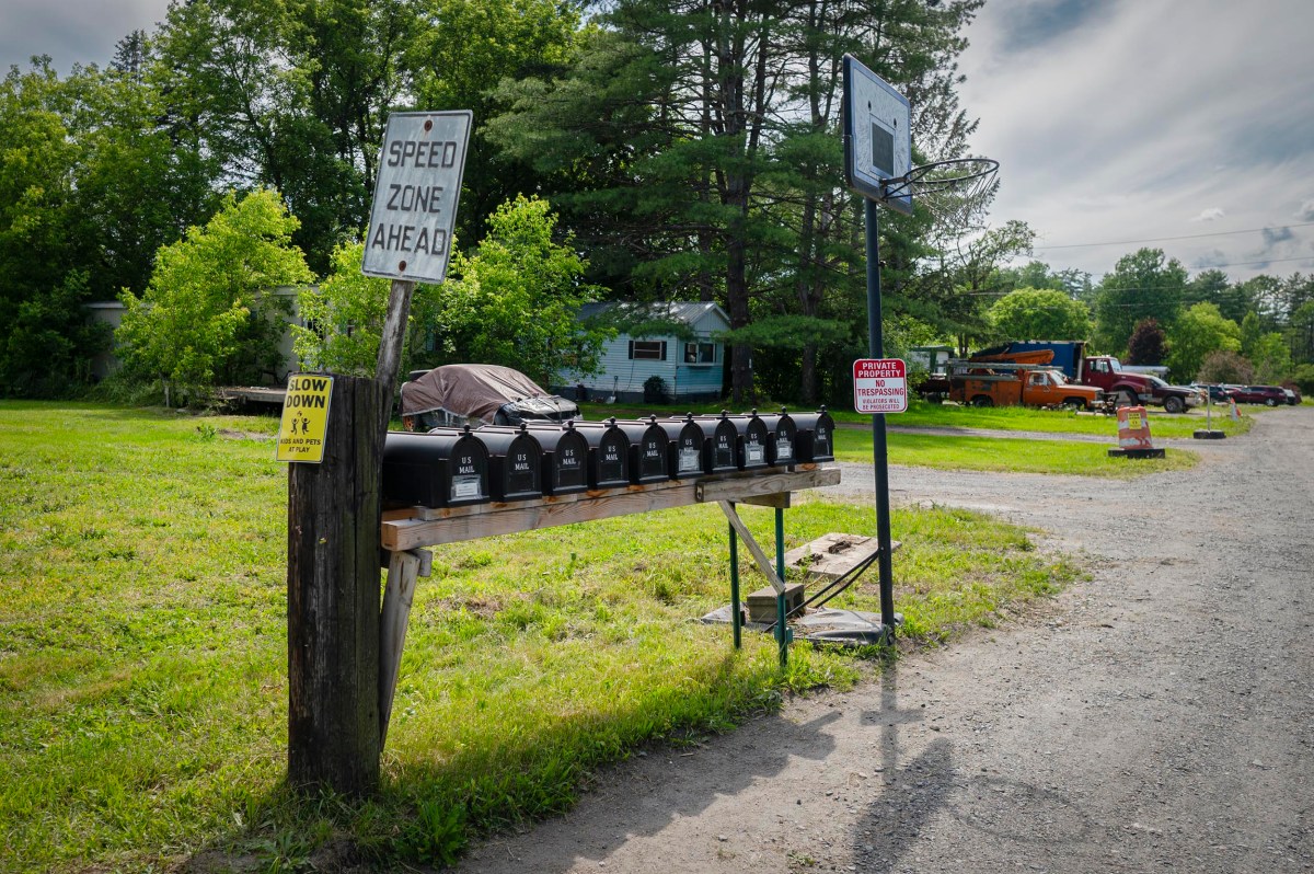 A row of mailboxes and a basketball hoop stand beside a dirt road, with a "Speed Zone Ahead" sign and trees in the background.