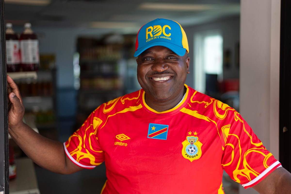 Smiling man wearing a red football jersey and a blue-yellow RDC cap stands in a shop doorway.
