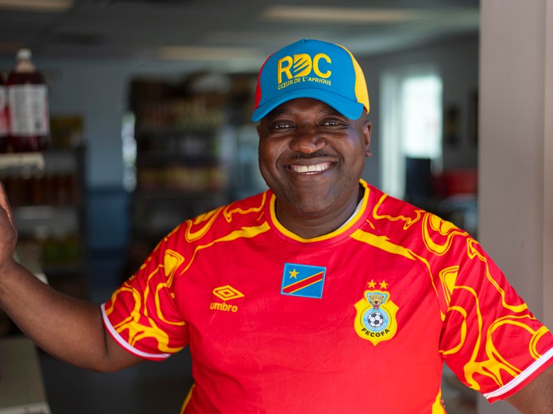 Smiling man wearing a red football jersey and a blue-yellow RDC cap stands in a shop doorway.