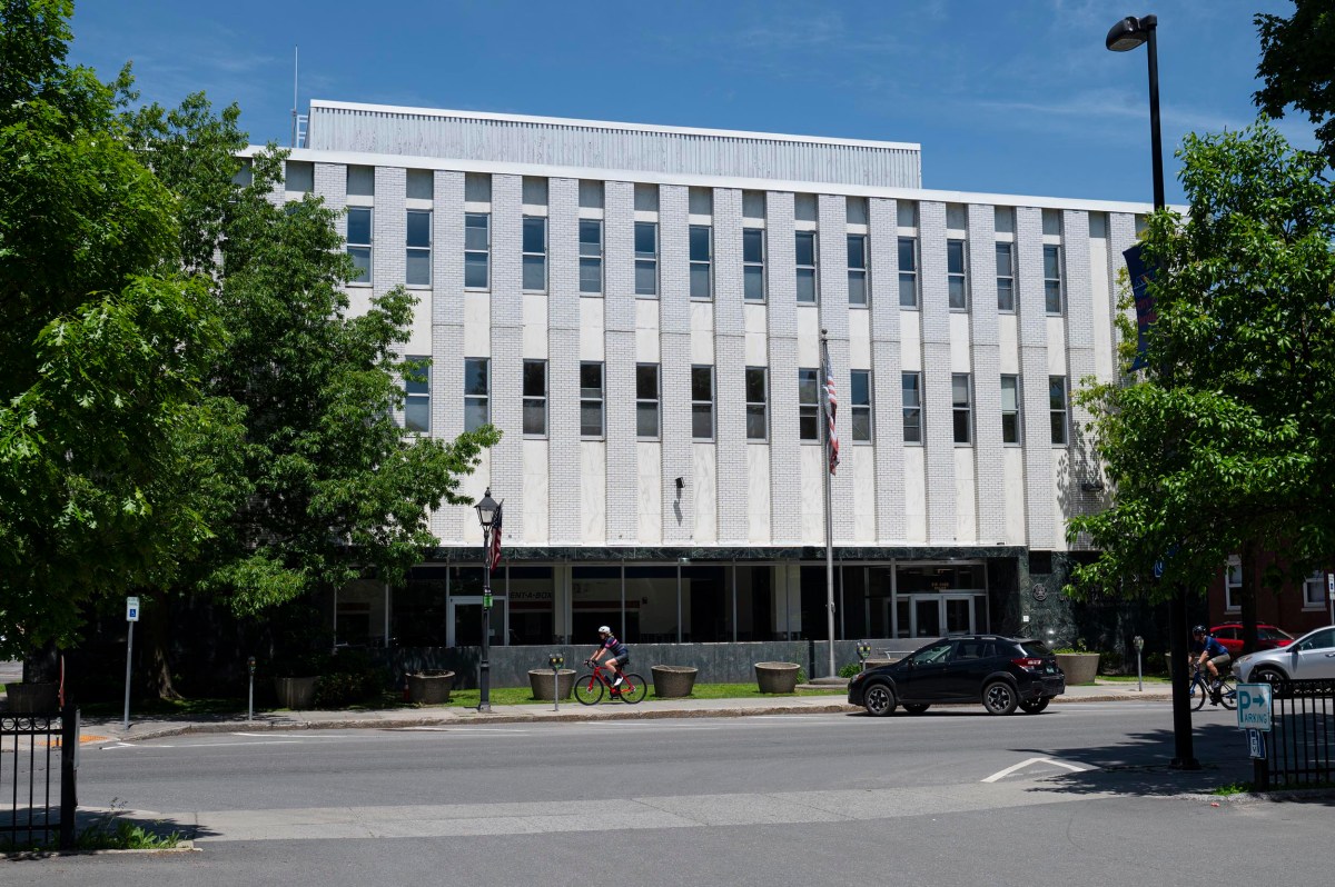 A person rides a bicycle past a three-story white office building with rows of windows and several flags at the entrance on a sunny day.