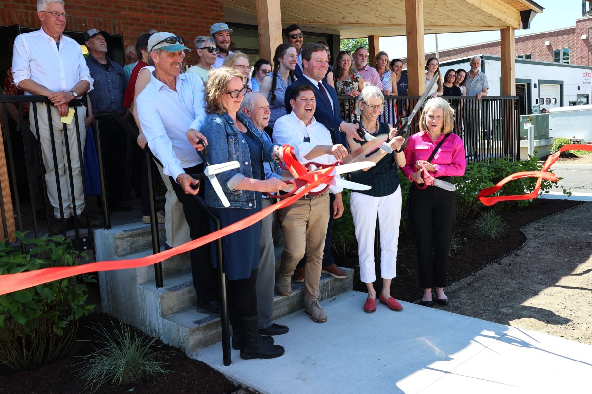 A group of people stands outside a building as several individuals cut a red ribbon with large scissors during a ribbon-cutting ceremony.