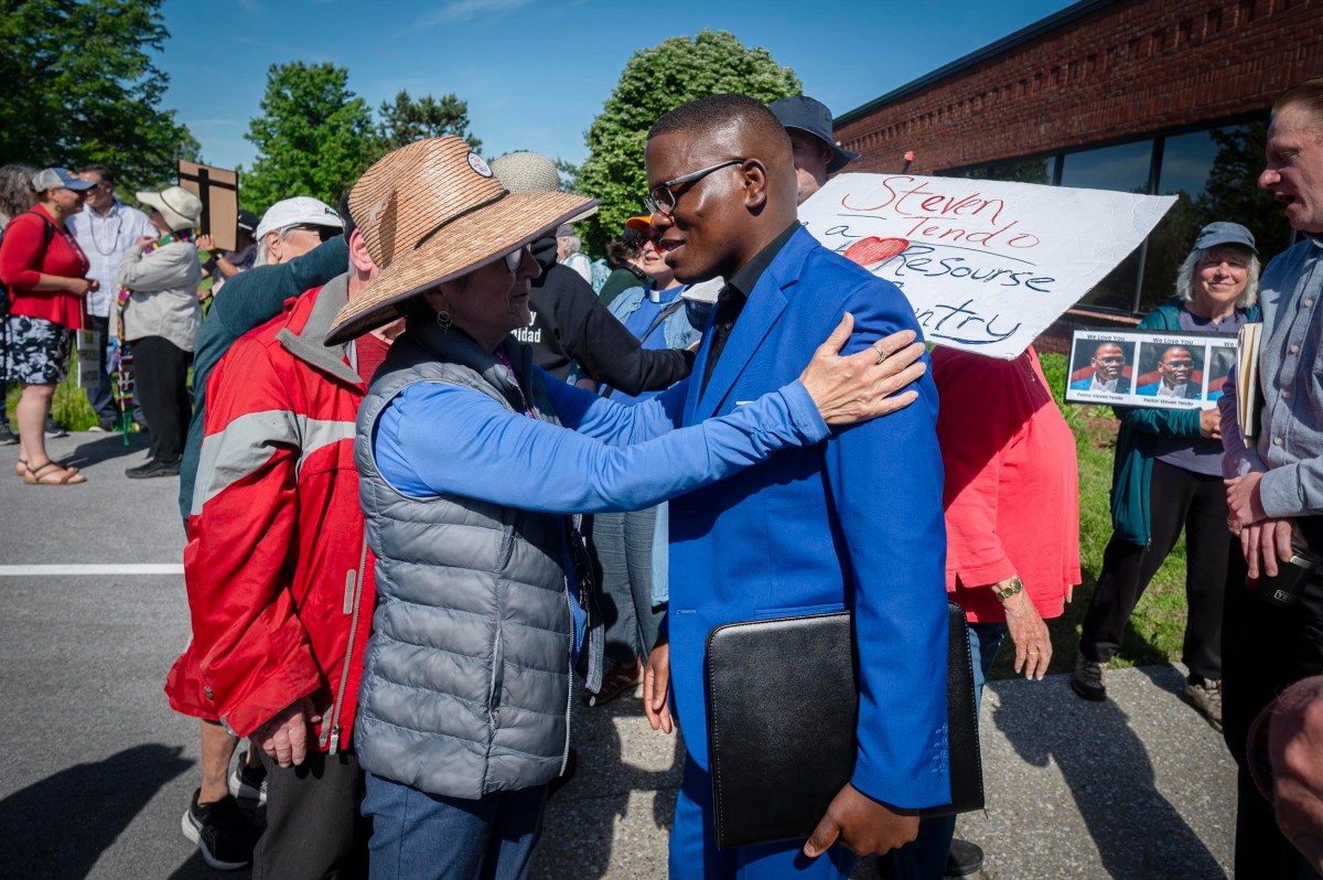 A woman in a hat embraces a man in a blue suit during an outdoor gathering, with people and a supportive sign in the background.
