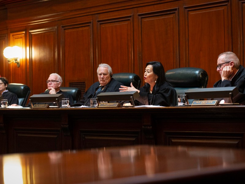 Five judges sit behind a wooden bench in a courtroom, with nameplates in front of them and an American flag visible on the left side.