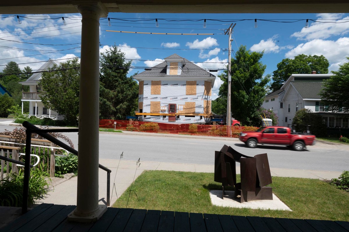 A house under construction is seen across the street, with a red pickup truck driving by and a metal sculpture on the lawn in the foreground.