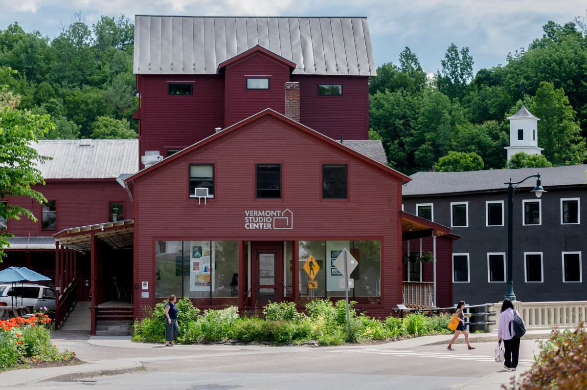 Red building with a sign that reads "Vermont Studio Center," surrounded by greenery, with people walking nearby and other buildings in the background.