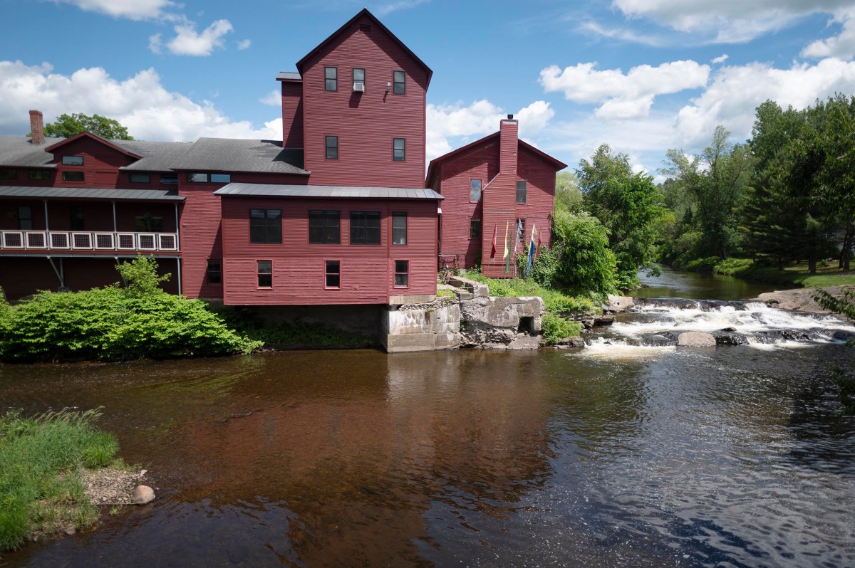 A large red building stands beside a small river and waterfall, surrounded by green trees under a partly cloudy sky.