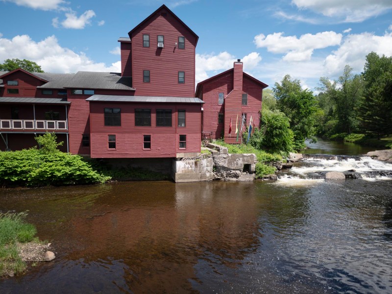 A large red building stands beside a small river and waterfall, surrounded by green trees under a partly cloudy sky.