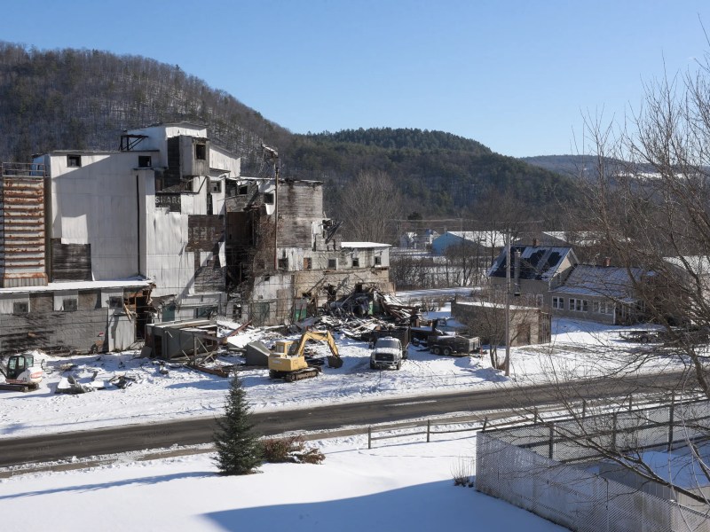 A partially destroyed industrial building with debris and construction equipment sits beside a snowy road in a small town, surrounded by hills and leafless trees.