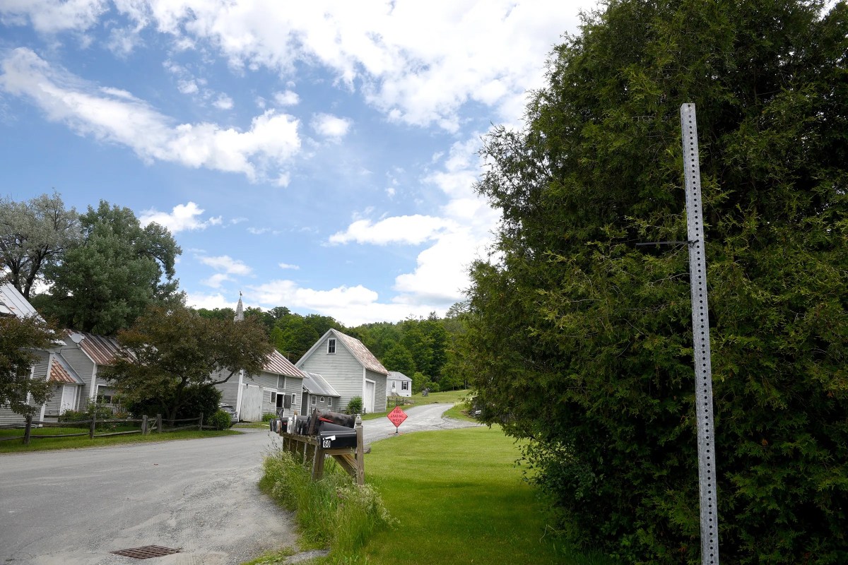 A rural road curves past a few white houses and mailboxes, bordered by grass, trees, and a blue sky with scattered clouds.