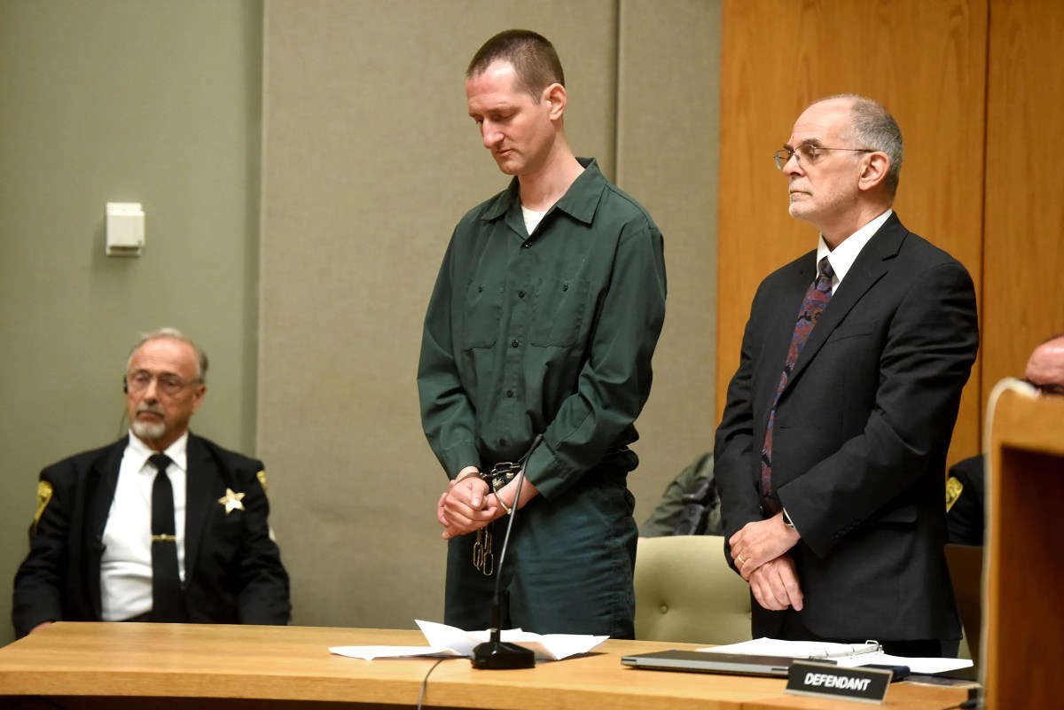 A man in handcuffs stands beside his attorney in a courtroom as a uniformed officer sits nearby; a "defendant" sign is visible on the table.