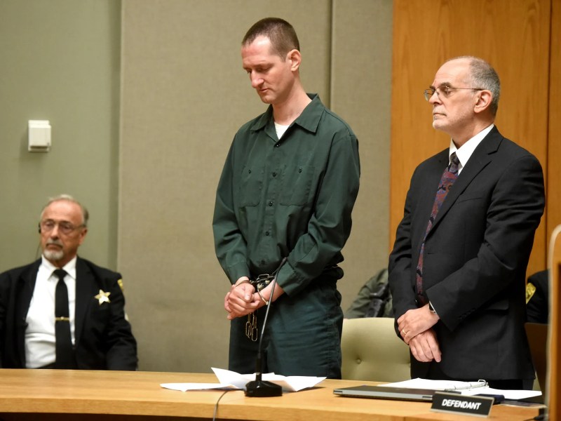 A man in handcuffs stands beside his attorney in a courtroom as a uniformed officer sits nearby; a "defendant" sign is visible on the table.