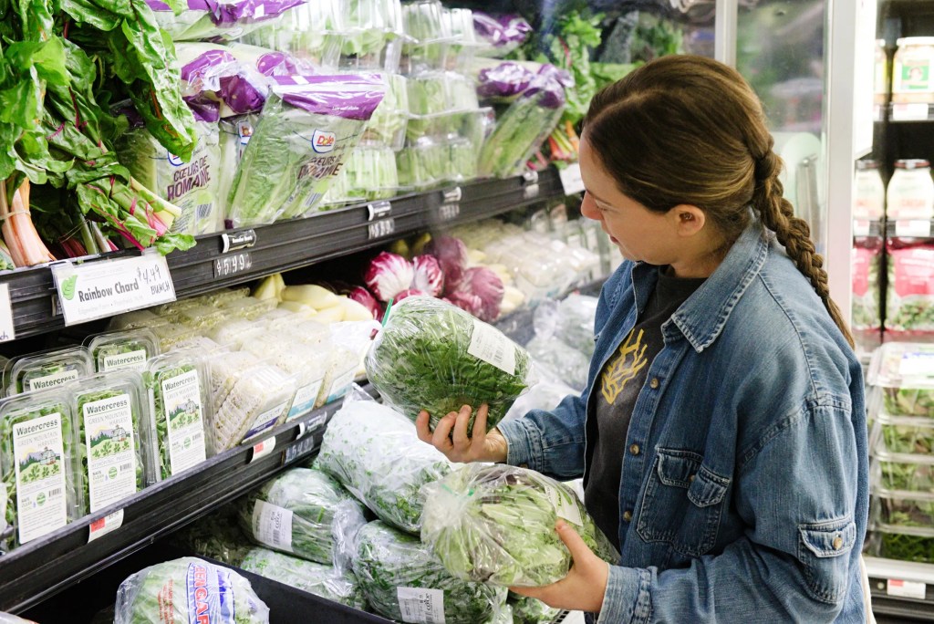 A person in a denim jacket examines packaged leafy greens in the produce section of a grocery store.