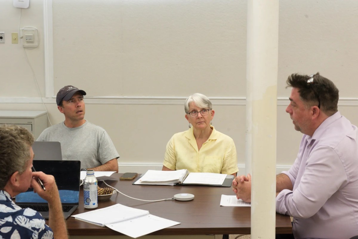 Four people are seated at a table having a discussion, with papers, a laptop, and a water bottle in front of them; one person is partially blocked by a pillar.