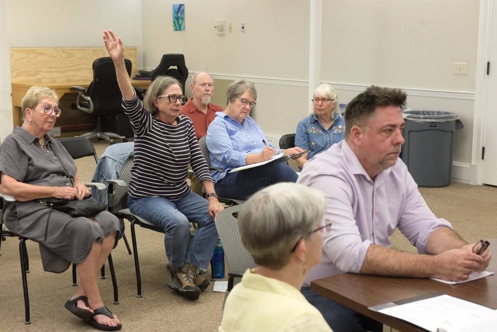 A woman raises her hand while sitting among others in a small meeting room; some people are taking notes, and one man sits at a table in the foreground.