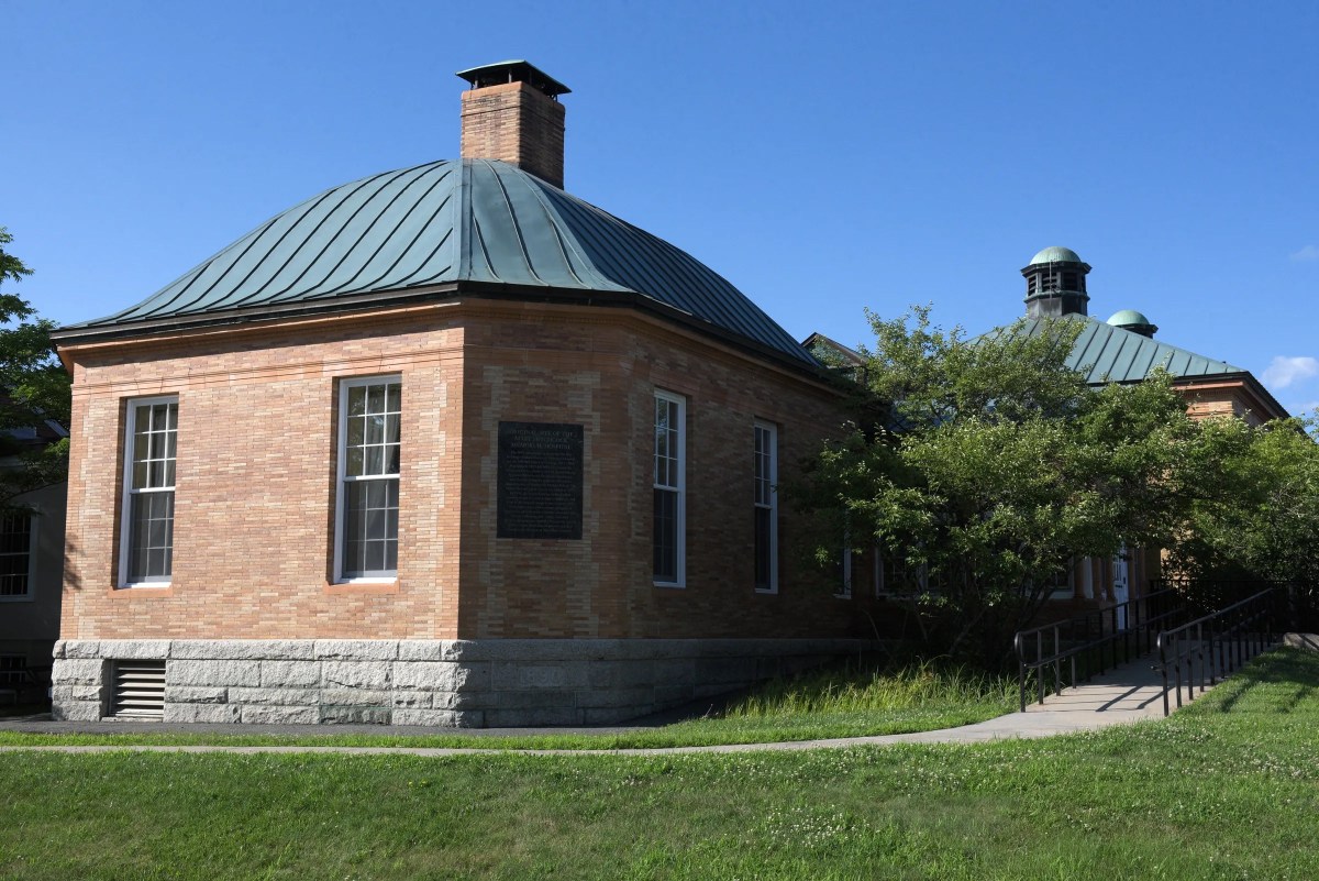 Brick building with green metal roof, large windows, and a plaque on the wall, surrounded by grass and trees under a clear blue sky.