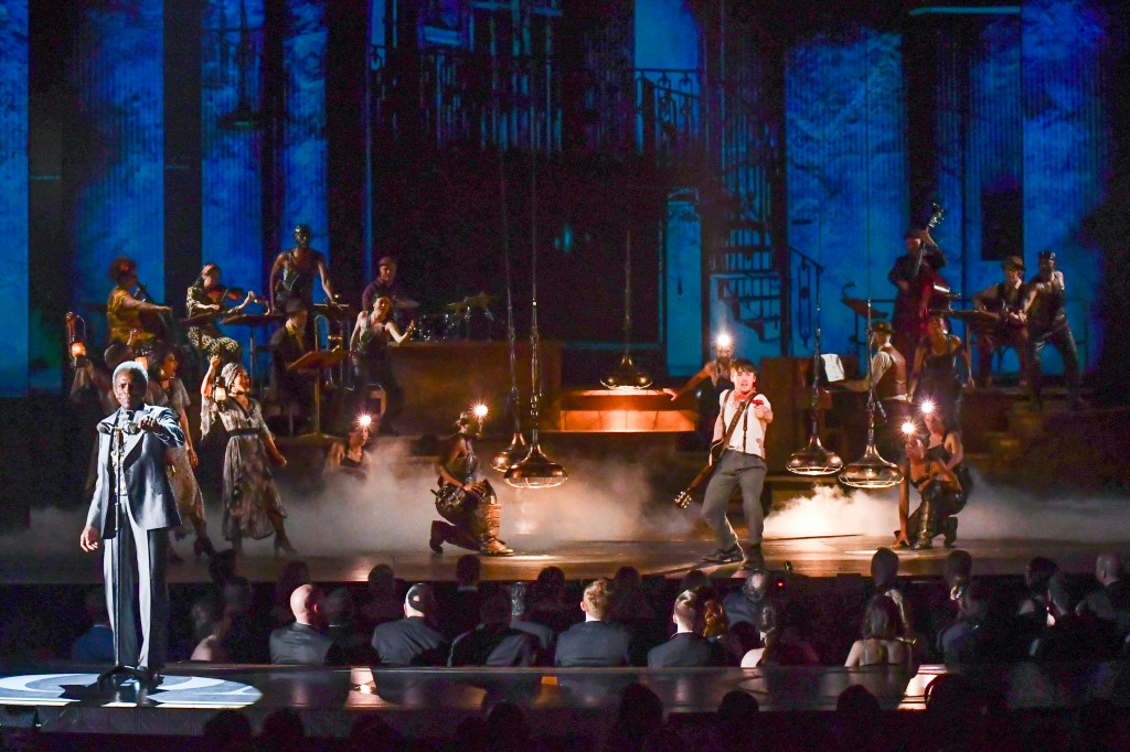 A live stage performance features a singer in the foreground, musicians playing various instruments, dramatic lighting, and a misty atmosphere with an audience watching.