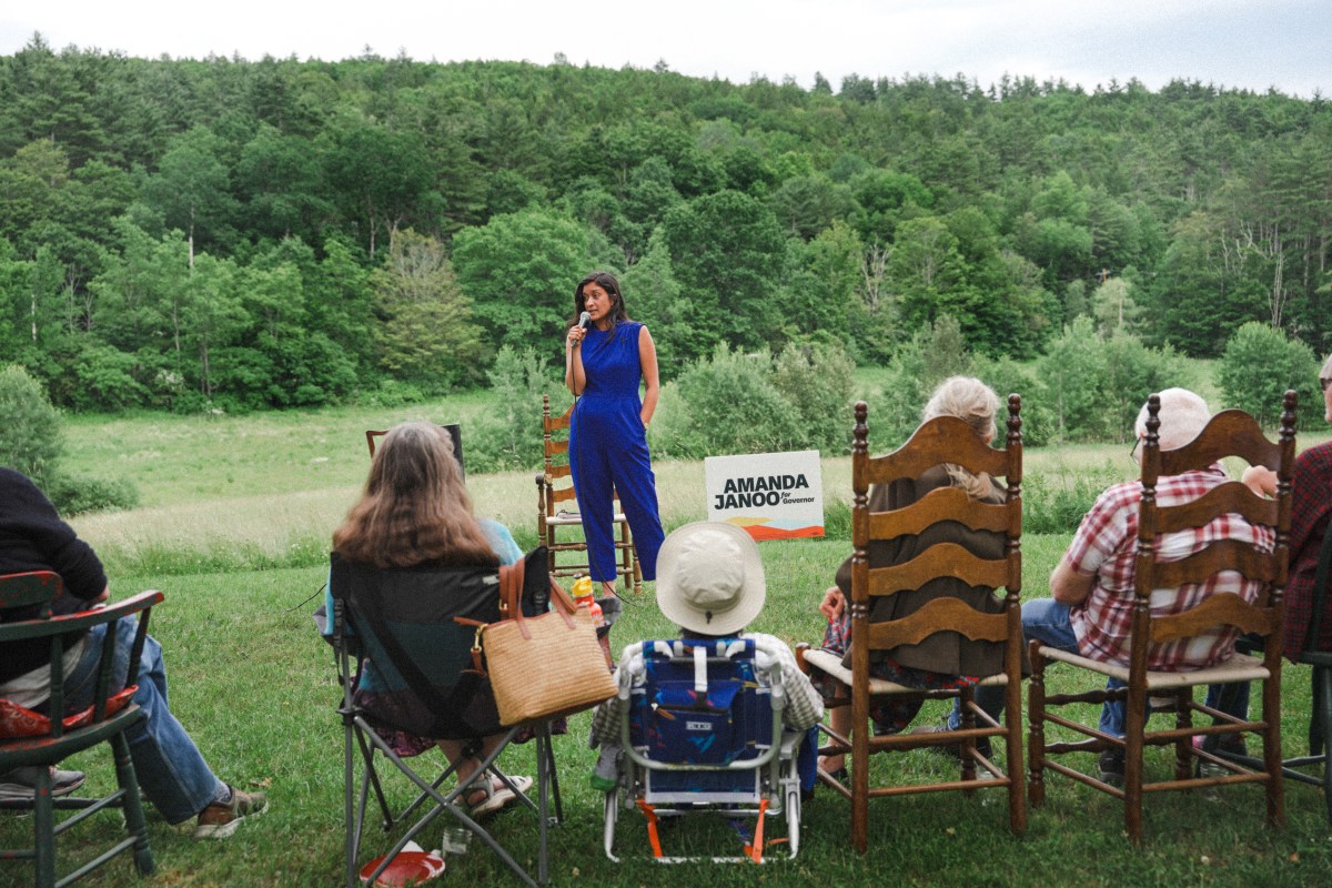 A woman in a blue outfit speaks into a microphone outdoors to a seated audience, with a sign reading "Amanda Janow" visible in the background. Trees and grass fill the landscape.
