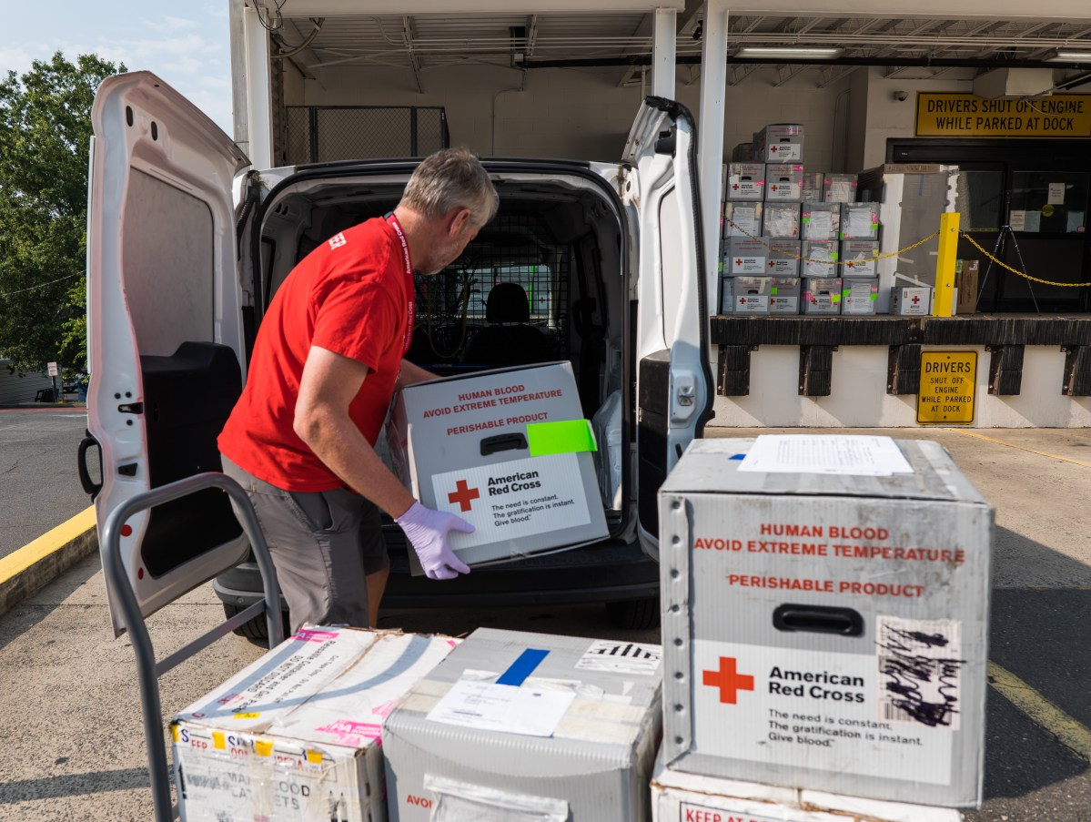 A person unloads boxes labeled "Human Blood, American Red Cross" from a vehicle at a delivery dock. Other labeled boxes are stacked nearby.
