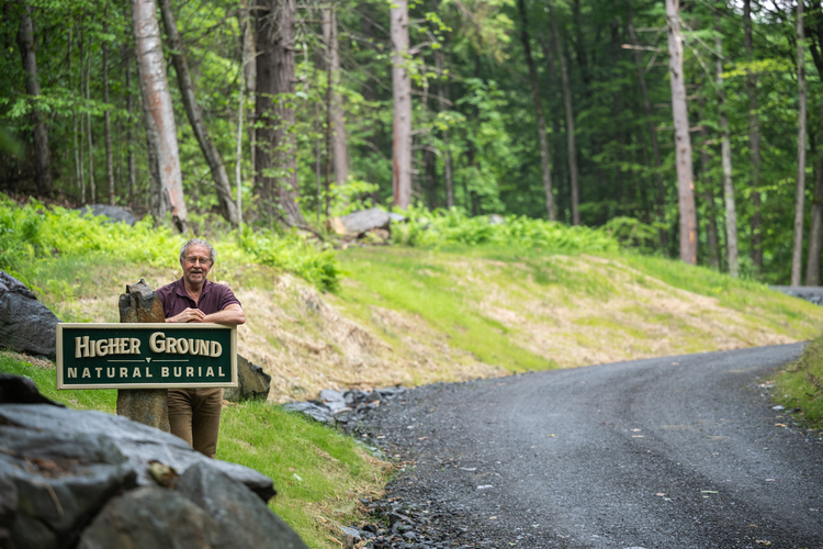 A person stands by a "Higher Ground Natural Burial" sign next to a gravel path in a wooded area.