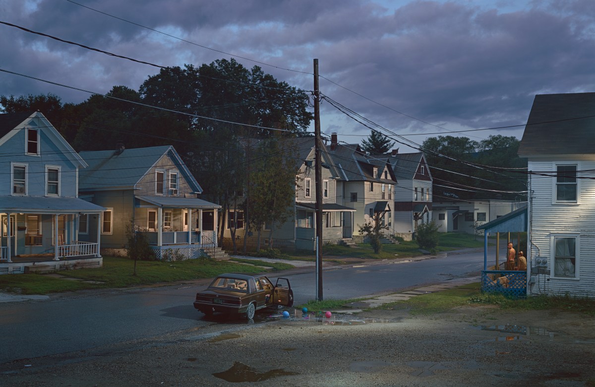 A quiet residential street at dusk with several houses, a parked car with open doors, and a person sitting on a porch lit by a warm light.