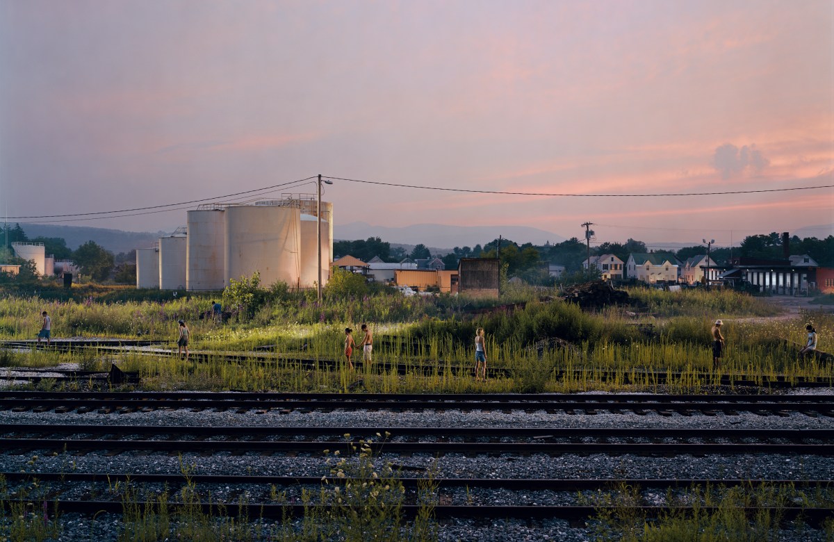 Several people walk and stand in overgrown grass near train tracks and large storage tanks at dusk, with houses and utility poles in the background.