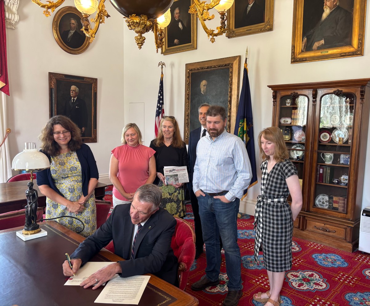 A man signs a document at a desk while five people stand behind him in a room with portraits, a display cabinet, and American flags.