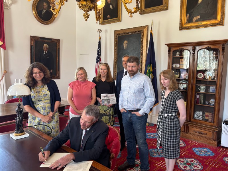 A man signs a document at a desk while five people stand behind him in a room with portraits, a display cabinet, and American flags.